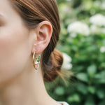 Gold hoop earrings with green enamel hummingbird and pink flower detail, elegant garden-inspired jewelry worn on model.