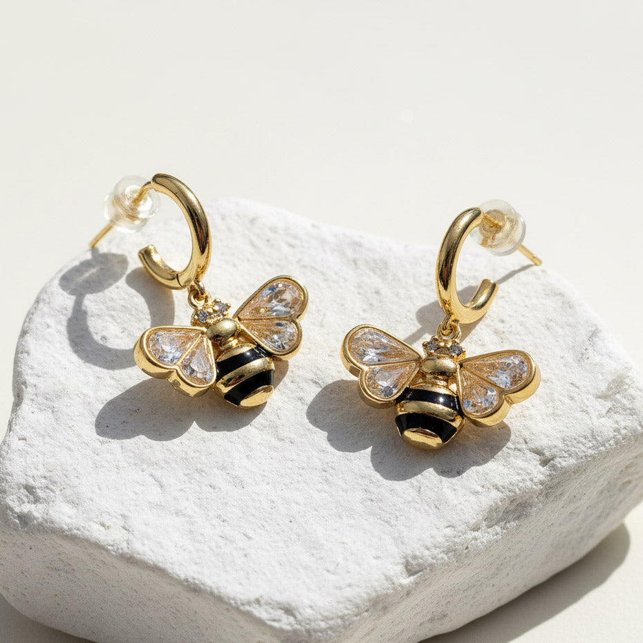 Gold bee-shaped earrings with black and clear gemstones on a white stone surface
