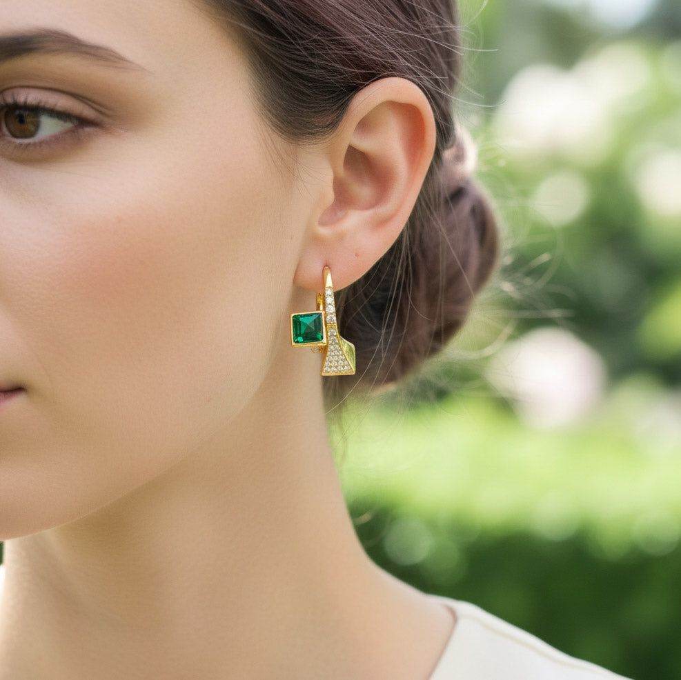 Close-up of a woman wearing gold earrings with green gemstones against a blurred natural background.