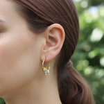 Close-up of a person wearing gold hoop earrings with a yellow gemstone against a blurred green background