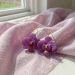 Purple orchid-shaped earrings on a pink fabric with a window in the background