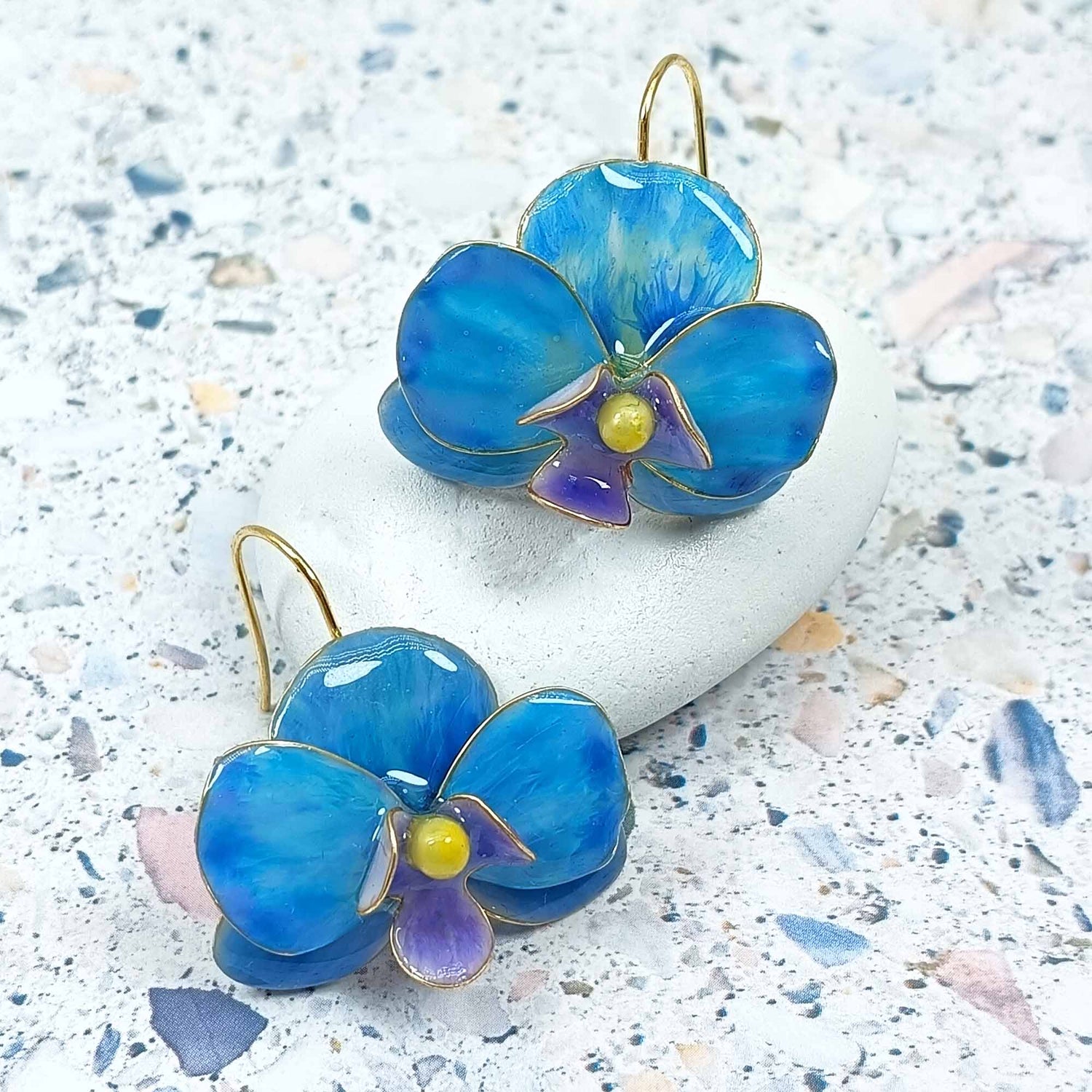Handcrafted blue enamel orchid earrings with gold-plated hooks, featuring detailed purple and yellow flower centers, displayed on a textured stone background.