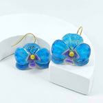 Pair of blue enamel orchid earrings with gold-plated hooks, featuring purple and yellow flower centers, displayed on a white curved stand.