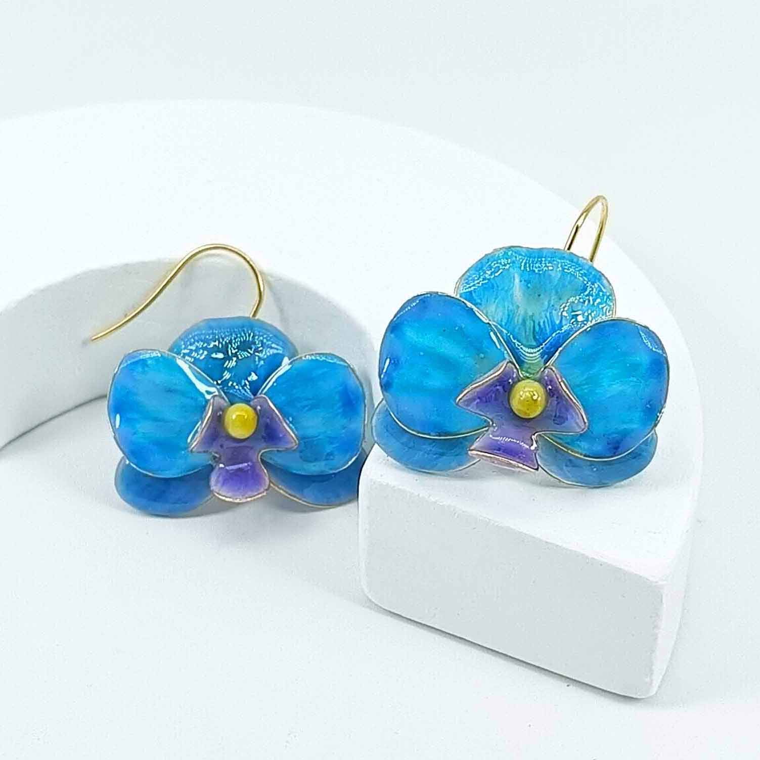 Pair of blue enamel orchid earrings with gold-plated hooks, featuring purple and yellow flower centers, displayed on a white curved stand.