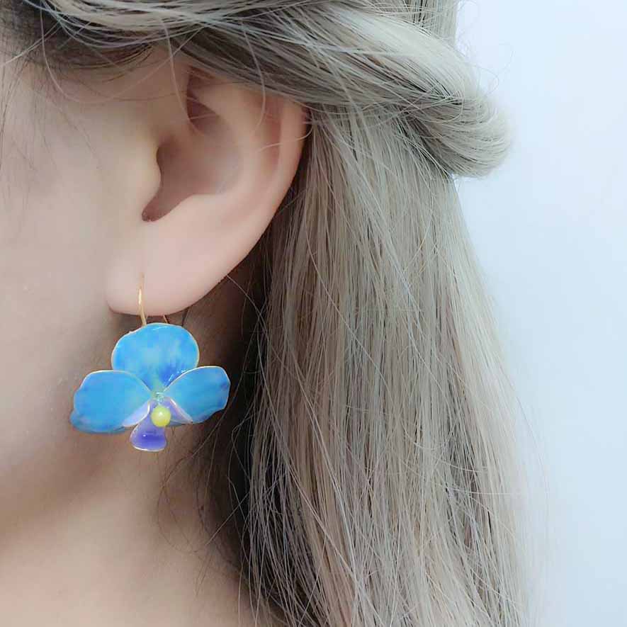 Model wearing a blue enamel orchid earring with gold hook, featuring a yellow center and purple petal detail, against a light background.