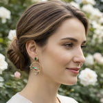 Woman wearing gold hoop earrings with green accents in a garden setting