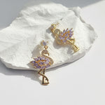 Close-up of gold flamingo drop earrings with purple CZ stones on textured white surface