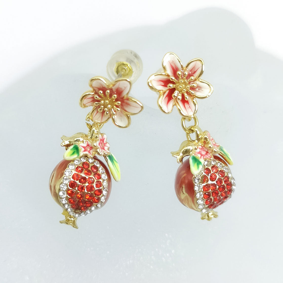 Pair of hand-painted enamel pomegranate and flower earrings with red cubic zirconia and crystal accents, displayed on a frosted glass surface.