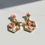 Hexagonal earrings with floral and bee designs on a light background