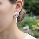 Close-up of a person wearing a floral earring with pearl drops against a blurred natural background.