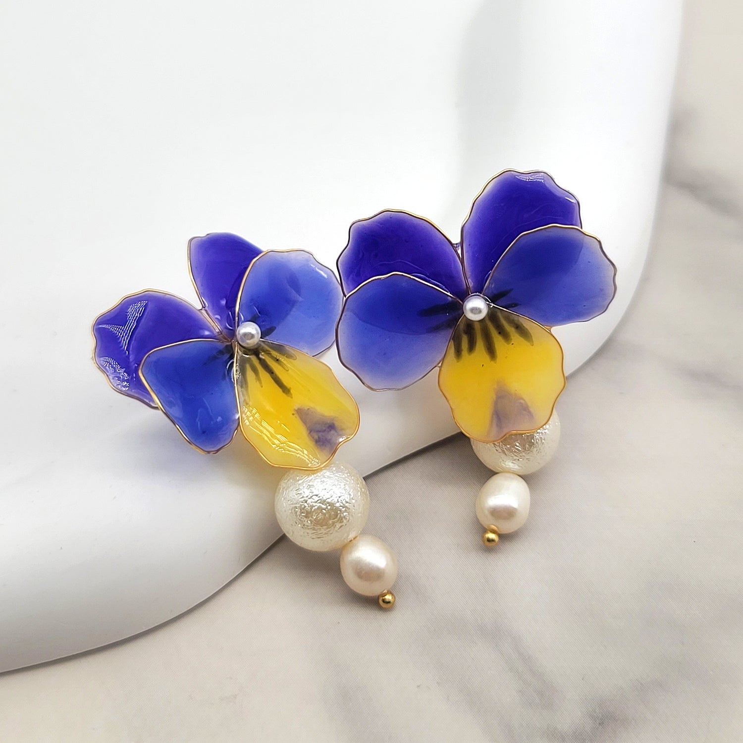 A pair of enamel floral earrings designed to resemble pansies with vibrant blue and yellow petals. Each earring features a small white pearl at the center of the flower and two larger pearls, one textured and one smooth, suspended below the floral motif. The edges of the petals are delicately outlined in gold, and the earrings are displayed on a white ceramic background.
