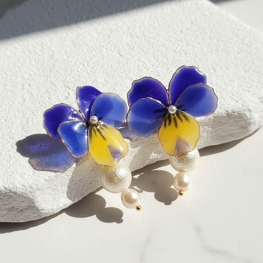 Hand-painted blue and yellow pansy flower earrings with pearl accents, displayed on a textured white surface.