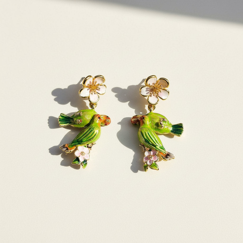 Hand-painted green parrot earrings with pink enamel flowers and gold detailing, displayed in sunlight