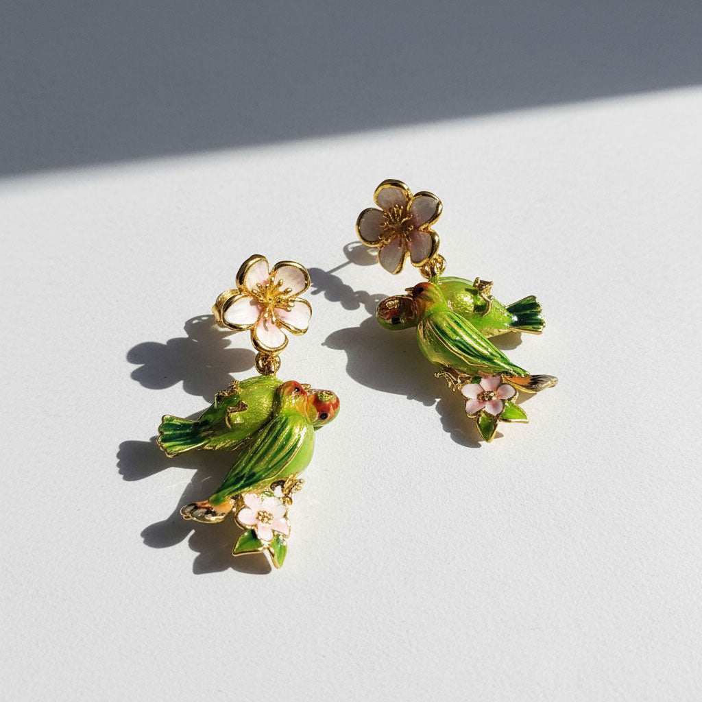 Gold earrings with hand-painted green parrots and pink enamel flowers, captured in bright natural light
