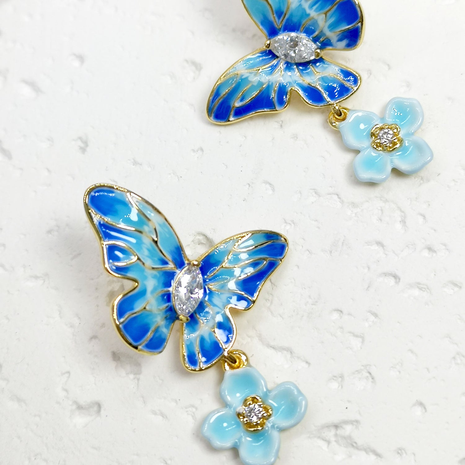 Close-up of blue enamel butterfly stud earrings with sparkling rhinestone centers and dangling light blue enamel flowers with crystal accents, set against a textured white background.