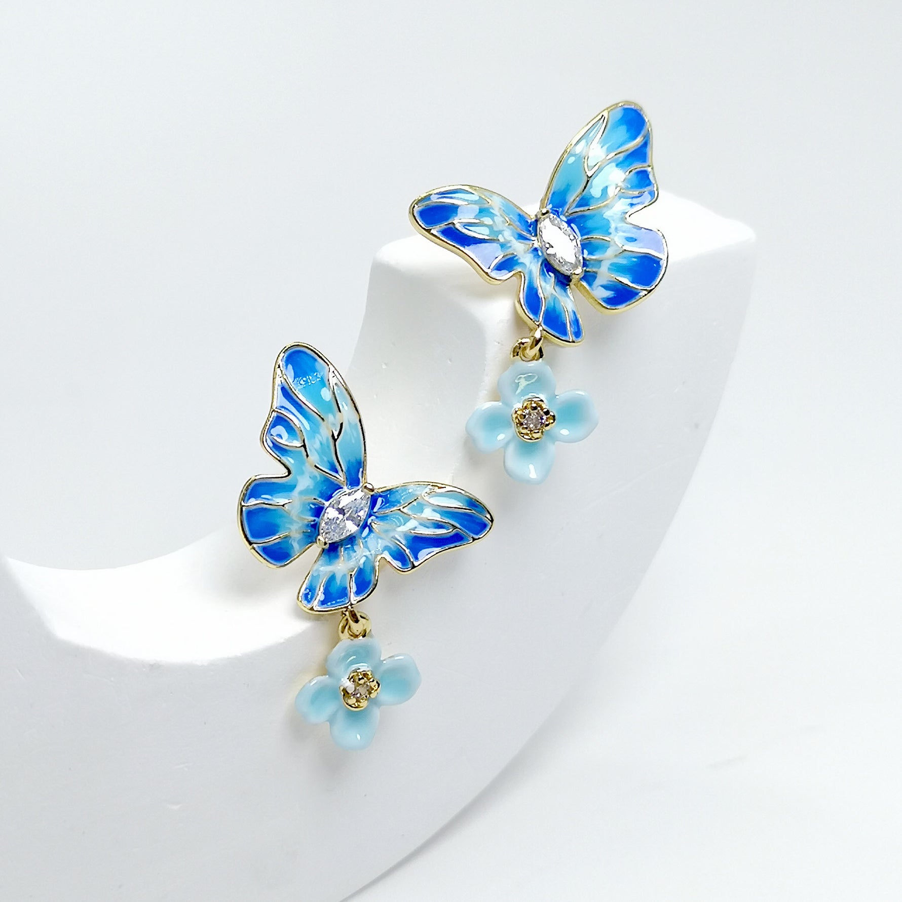 A pair of gold-plated stud earrings featuring hand-painted blue enamel butterflies with sparkling cubic zirconia centers and dangling pastel blue flower charms.