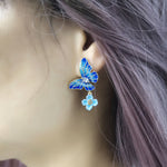 A close-up of a person wearing a blue enamel butterfly earring with a small dangling light blue flower charm, accented with crystal details. The earring is worn on the left ear, framed by lavender-toned hair.
