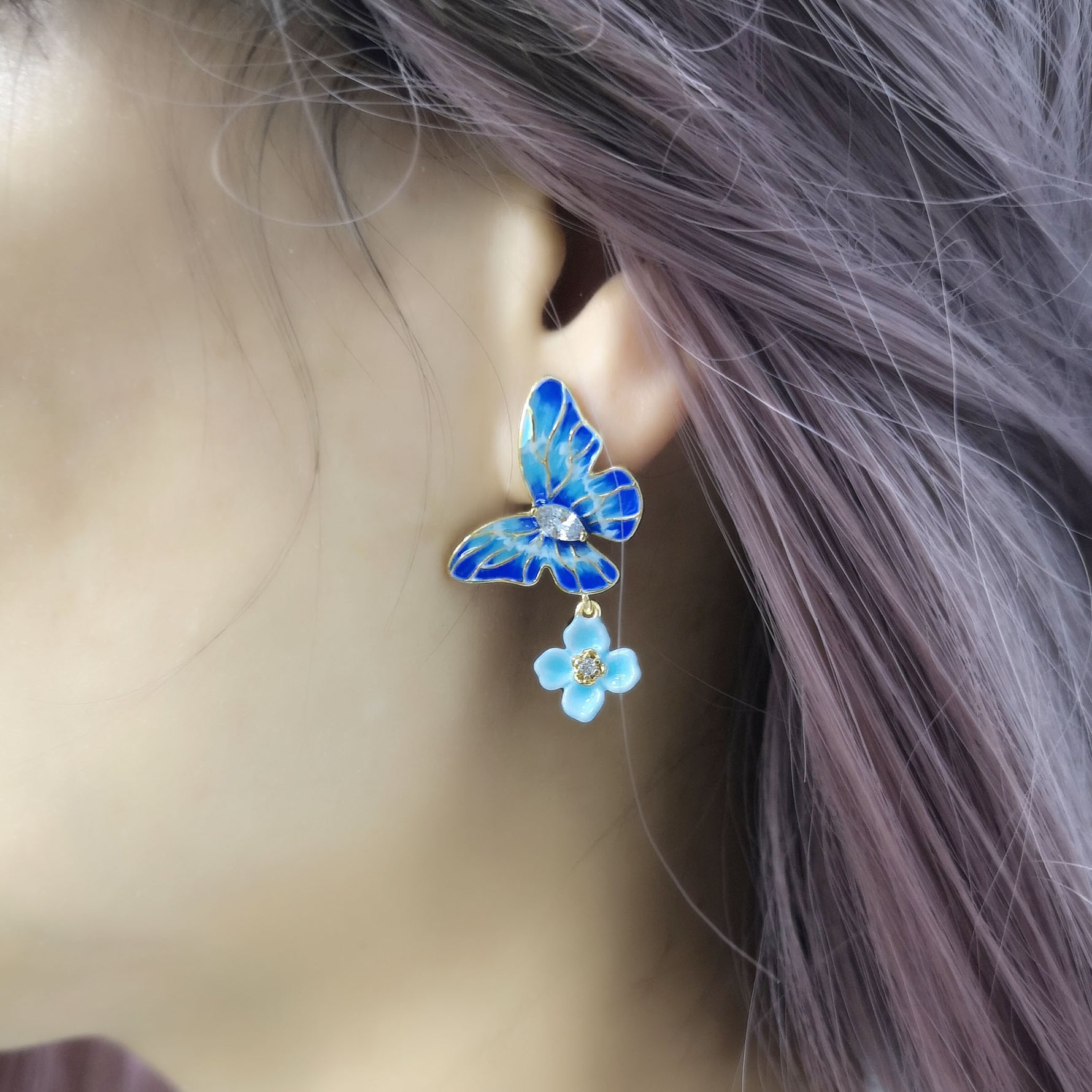A close-up of a person wearing a blue enamel butterfly earring with a small dangling light blue flower charm, accented with crystal details. The earring is worn on the left ear, framed by lavender-toned hair.