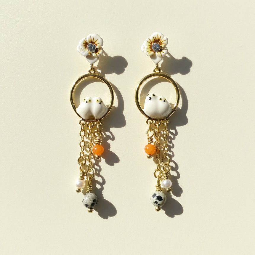 Pair of earrings with floral tops, bird-like figures, and hanging beads on a beige background