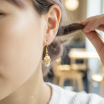 Close-up of a person wearing a gold earring with a small figure, blurred indoor background