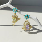 Frog-shaped drop earrings hanging from turquoise flower studs in sunlight.
