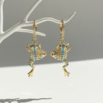 Gold frog-shaped earrings with blue gemstones on a white stand against a gray background