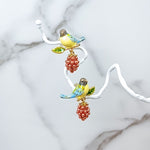 Close-up of hand-painted bird and berry drop earrings, displayed on a white branch.