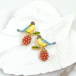 Pair of bird and berry stud earrings with blue and yellow hand-painted enamel birds and clusters of small red beads, resting on a textured white background.
