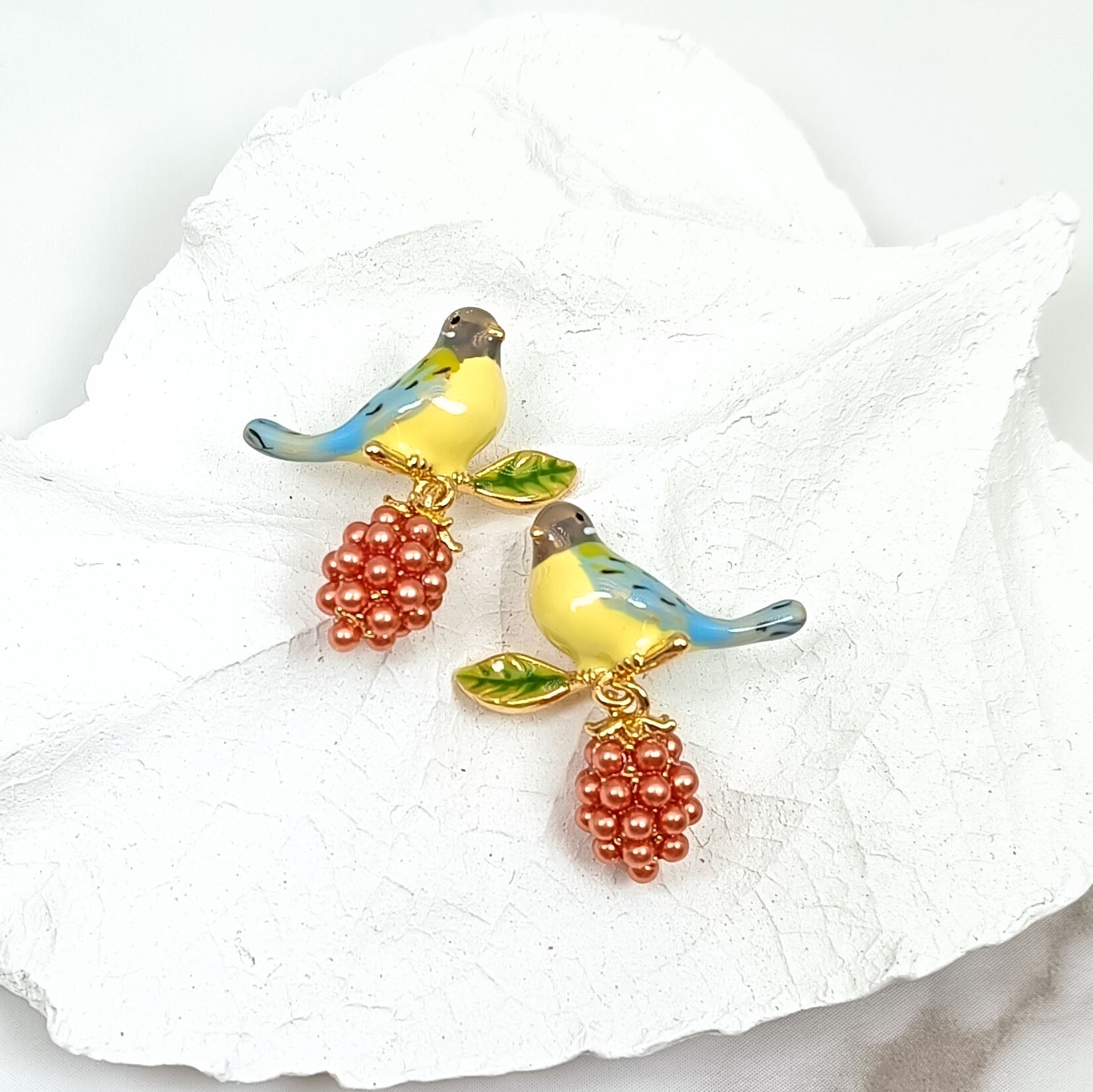Pair of bird and berry stud earrings with blue and yellow hand-painted enamel birds and clusters of small red beads, resting on a textured white background.