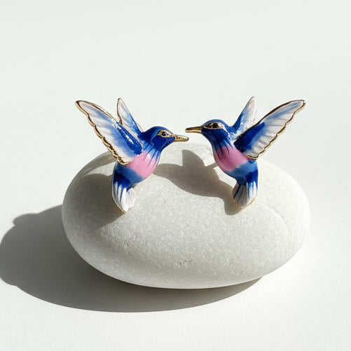 Close-up of blue and pink enamel hummingbird earrings displayed on white stone, natural light.