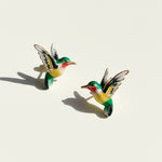 Green and yellow enamel hummingbird stud earrings with gold trim on light background, sunlight and shadow.