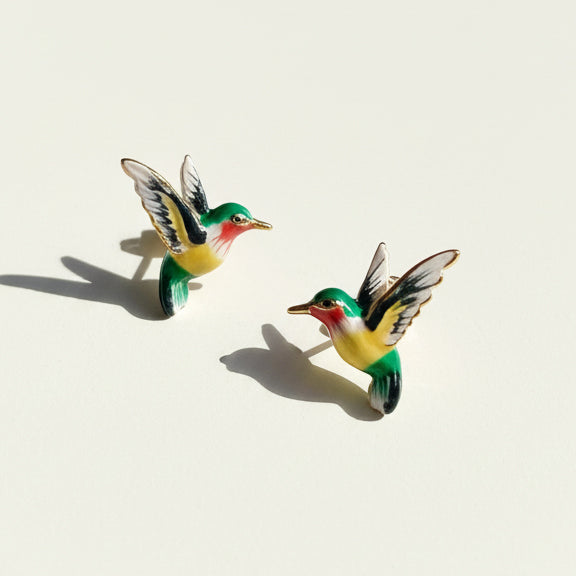 Green and yellow enamel hummingbird stud earrings with gold trim on light background, sunlight and shadow.