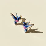 Blue and pink hand-painted enamel hummingbird stud earrings on cream background with sunlight and shadow.