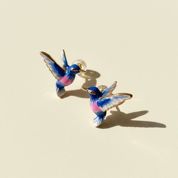 Blue and pink hand-painted enamel hummingbird stud earrings on cream background with sunlight and shadow.