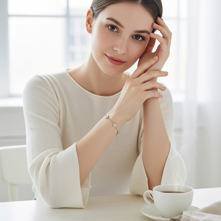 Woman in a white sweater sitting at a table with a cup of coffee wearing a moissanite flower chain bracelet, smiling.