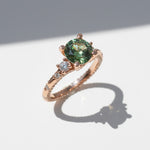 Rose gold ring with a round green moissanite center stone and side stones on white background.