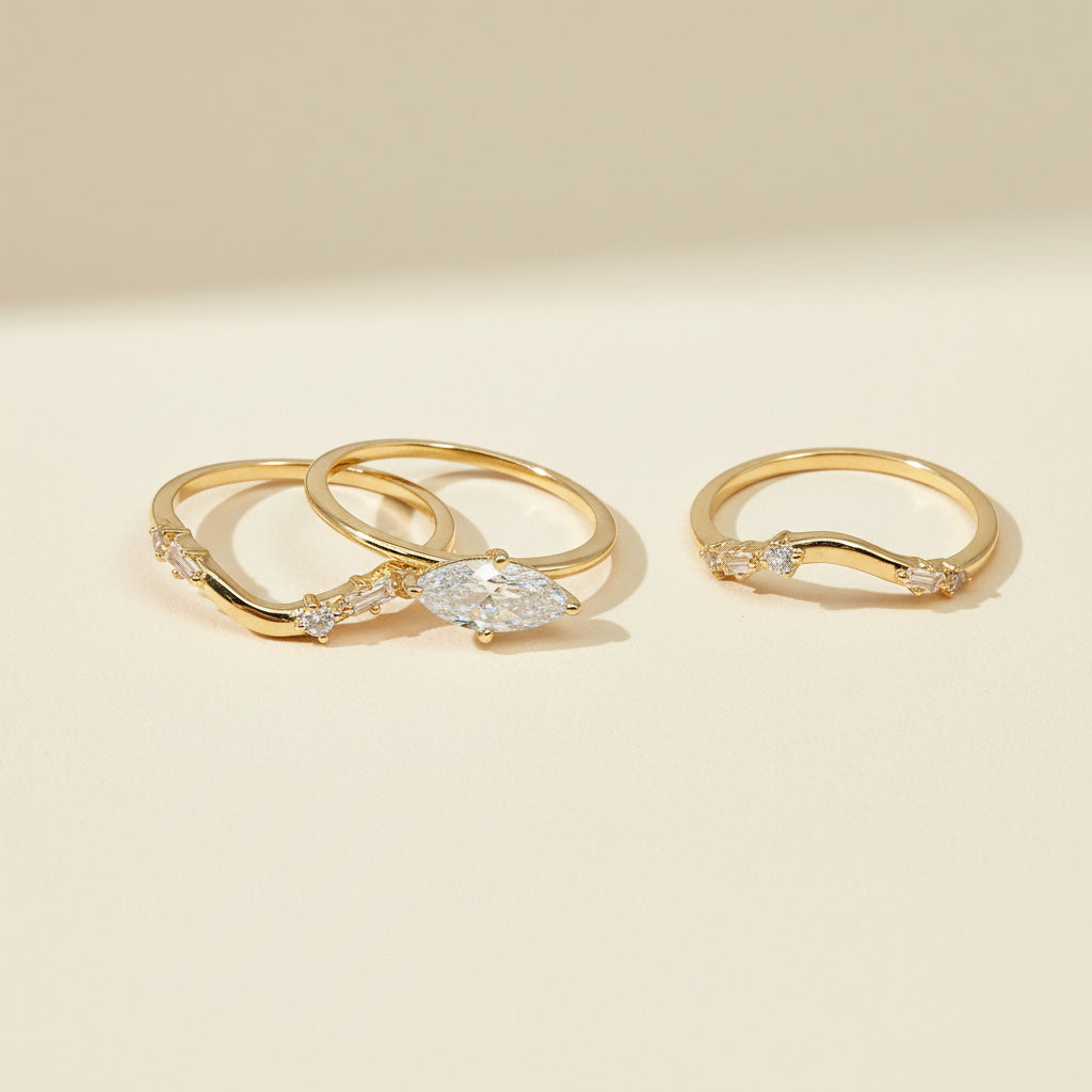 A set of three gold-plated silver rings displayed on a light beige surface. The set includes one ring with a marquise-cut moissanite center stone and two curved bands adorned with round and baguette-cut accent stones. The minimalist, elegant design allows the rings to be worn stacked or separately for a refined look.