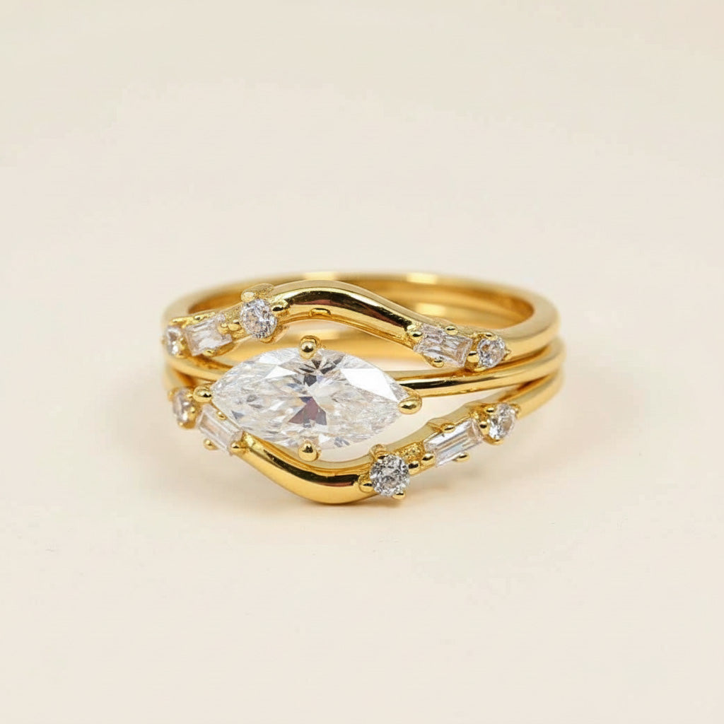 A set of three gold-plated silver rings featuring a marquise-cut moissanite center stone and two matching curved bands embellished with round and baguette-cut accent stones. The stacked design forms an elegant, symmetrical look with brilliant sparkle against a soft beige background.