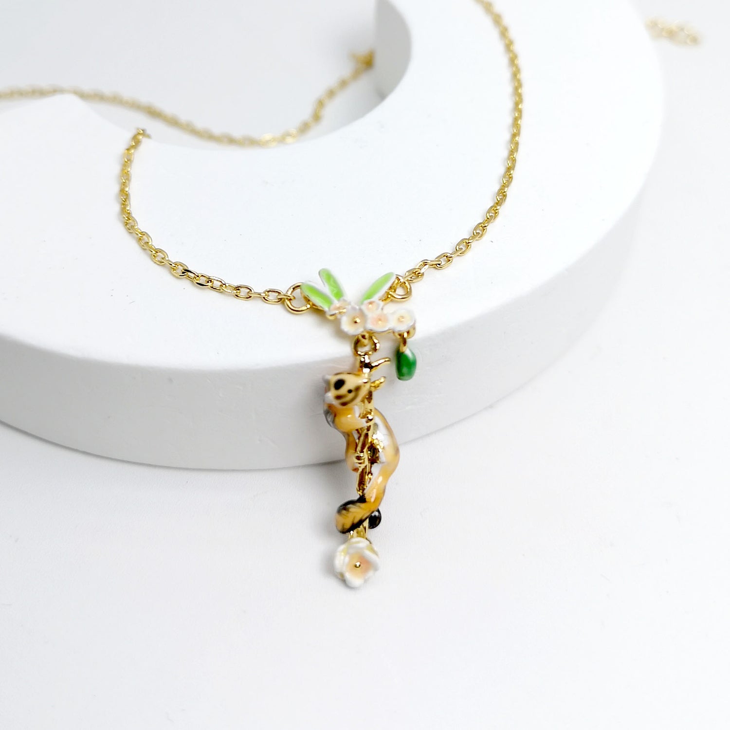A gold-plated necklace featuring a whimsical pendant of a hand-painted enamel chipmunk climbing a vine adorned with delicate white flowers and green leaves. The pendant hangs from a fine gold chain and is displayed on a white, curved stand to showcase its intricate, playful design.