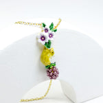 Close-up of a delicate pendant necklace featuring a yellow bird, white and purple flowers, and a bunch of purple grapes, all crafted with hand-painted enamel on a gold-plated chain.