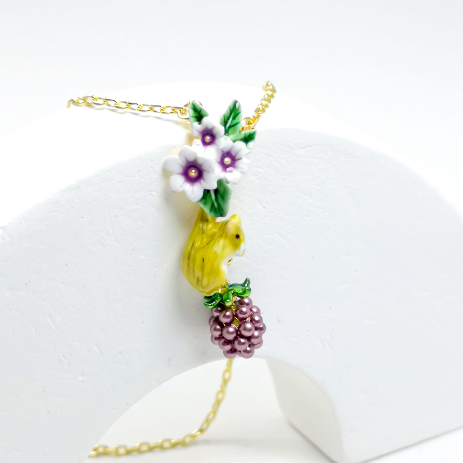 Close-up of a delicate pendant necklace featuring a yellow bird, white and purple flowers, and a bunch of purple grapes, all crafted with hand-painted enamel on a gold-plated chain.