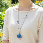 Person wearing a necklace with a blue flower pendant in a garden setting