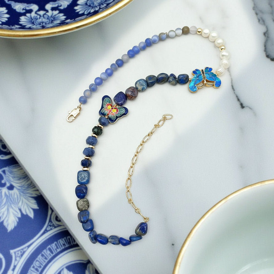 Handcrafted necklace with blue gemstone beads, enamel butterfly charms, and pearls on marble background.