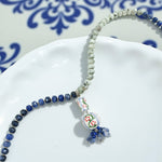 Handcrafted necklace with blue and white ceramic pendant, green and navy gemstone beads on white plate.