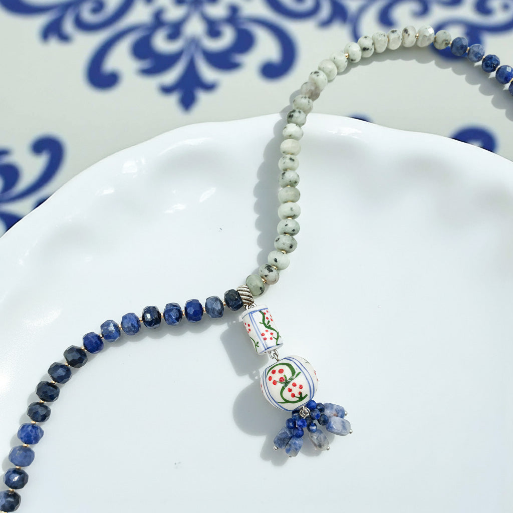 Handcrafted necklace with blue and white ceramic pendant, green and navy gemstone beads on white plate.