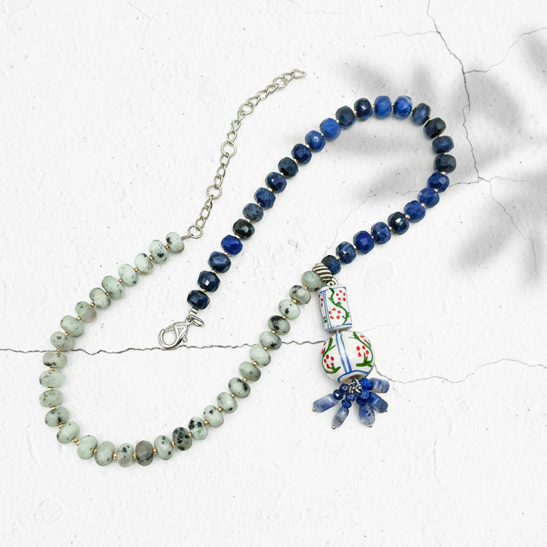 Beaded necklace with blue sodalite, green jade beads, and hand-painted ceramic pendant on textured background.