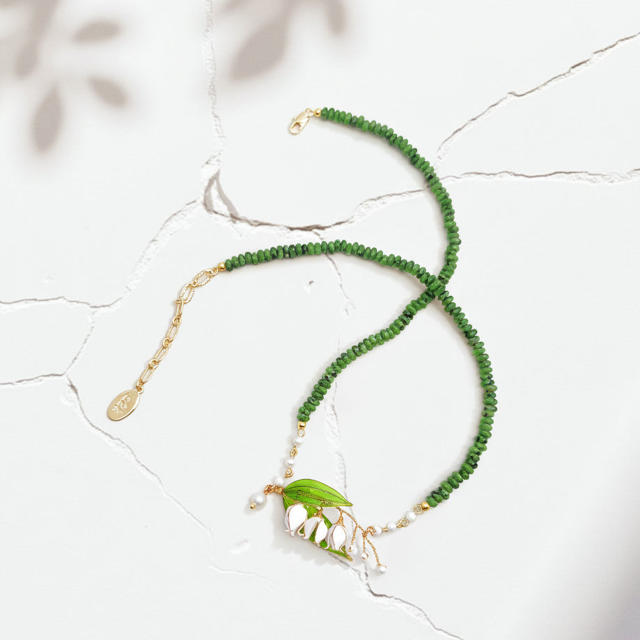 Green beaded necklace with enamel leaf and pearl pendant on cracked white background with sunlight and leaf shadows