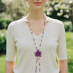 Woman wearing a long beaded necklace with a purple flower pendant outdoors.