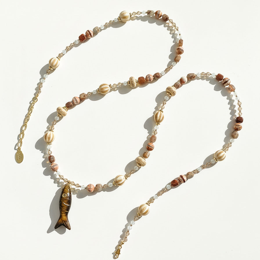 Long gemstone and ceramic bead necklace with carved fish pendant laid out in sunlight