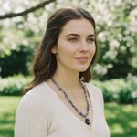 Woman wearing a blue beaded necklace outdoors with greenery in the background